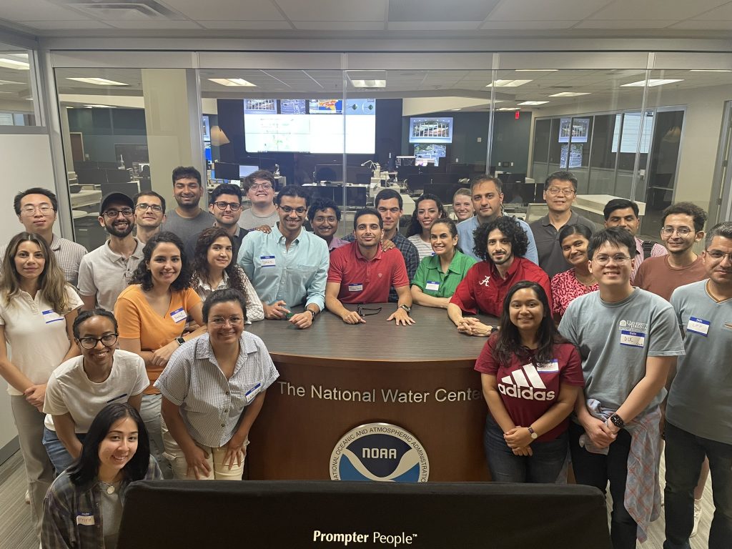 A group of people posing for a photo inside the National Water Center. They are participants of the CIROH National Water Center Innovators Program Summer Institute. The group includes both men and women, with diverse backgrounds, and they are dressed in casual attire. The background shows a control room with multiple monitors and workstations. The NOAA logo is prominently displayed on the desk in front of the group.