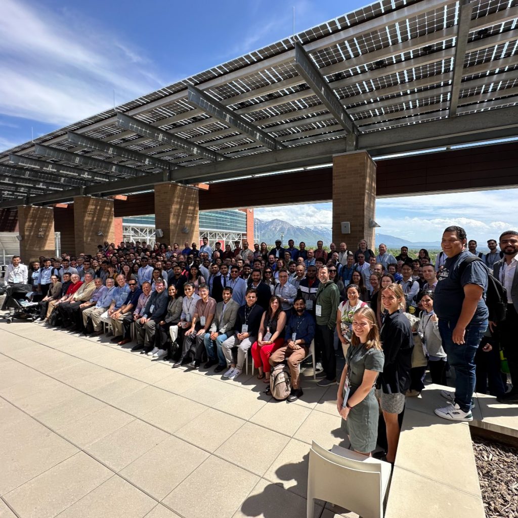 A large group of people gathered for a group photo under a covered outdoor area. They are attending the CIROH Second Annual Developers Conference. The group includes a diverse mix of men and women, dressed in business casual attire, standing and sitting in several rows. In the background, there are mountains and a clear blue sky.