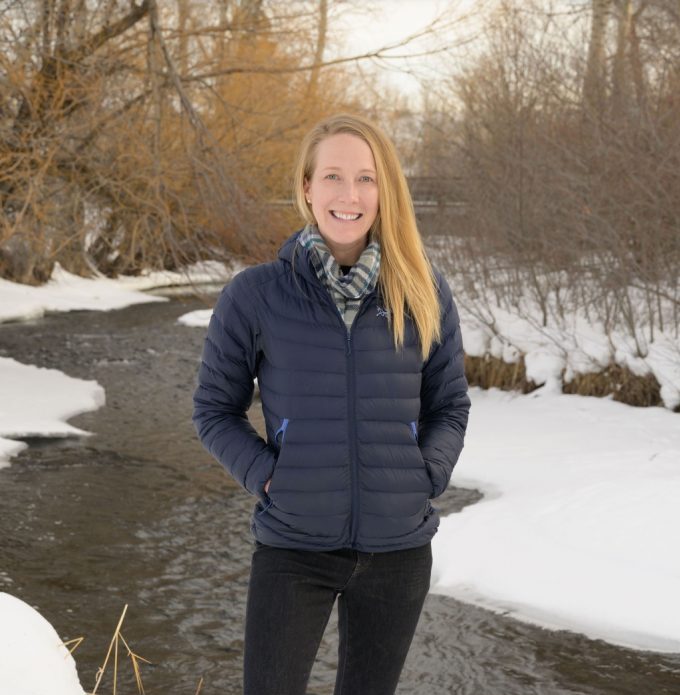 Person standing on snow beside a partially frozen stream, wearing a navy jacket, scarf, black jeans, and brown boots, with bare winter trees in the background.
